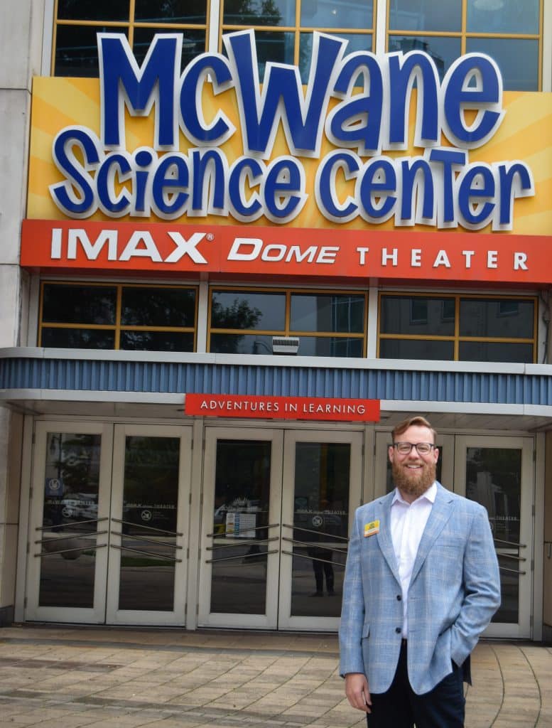 Brandan Lanman, new CEO of McWane Science Center, smiling and standing outside the center's entrance wearing a light blue plaid blazer and white shirt.