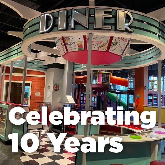 Retro-style diner play area inside Itty Bitty Magic City at McWane Science Center, with colorful play structures in the background and bold text reading 'Celebrating 10 Years.